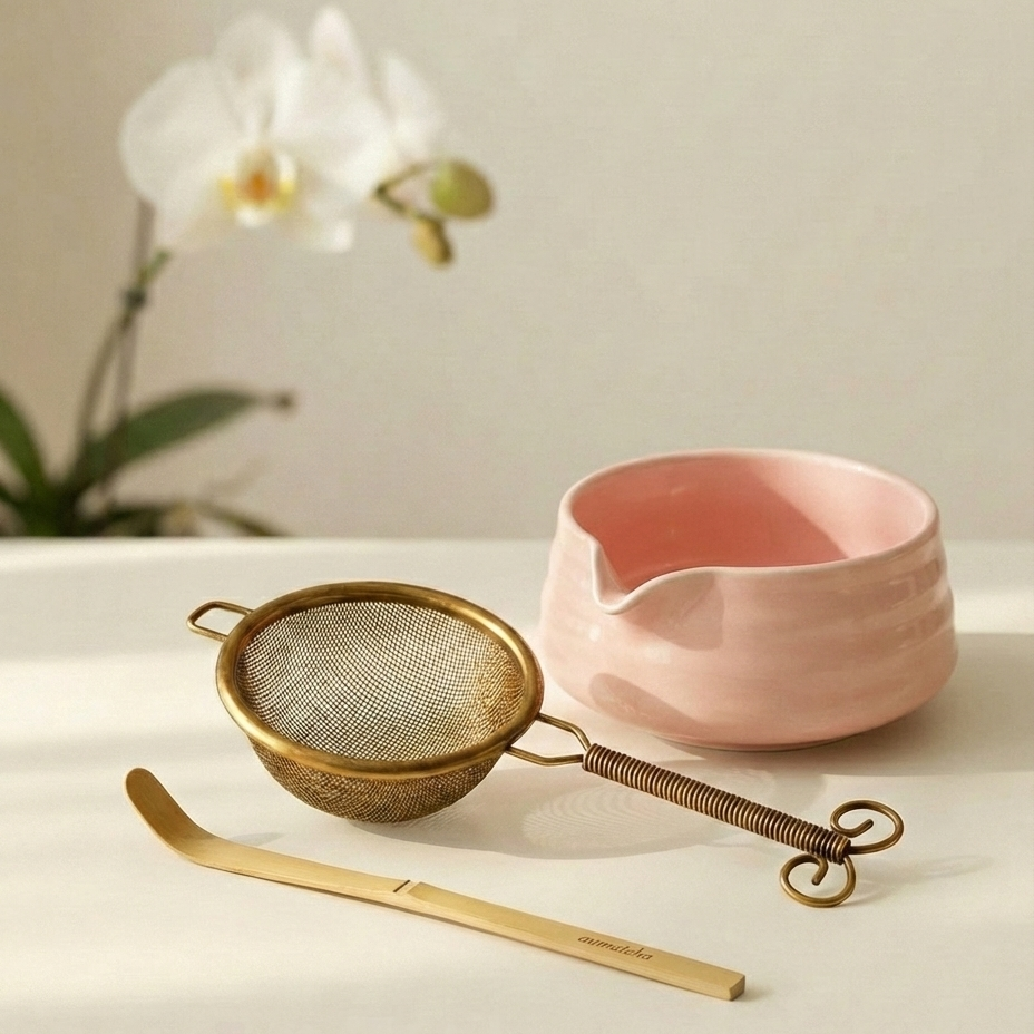 Gold matcha strainer and bamboo spoon with a pink ceramic bowl on a light surface, accompanied by a white orchid.