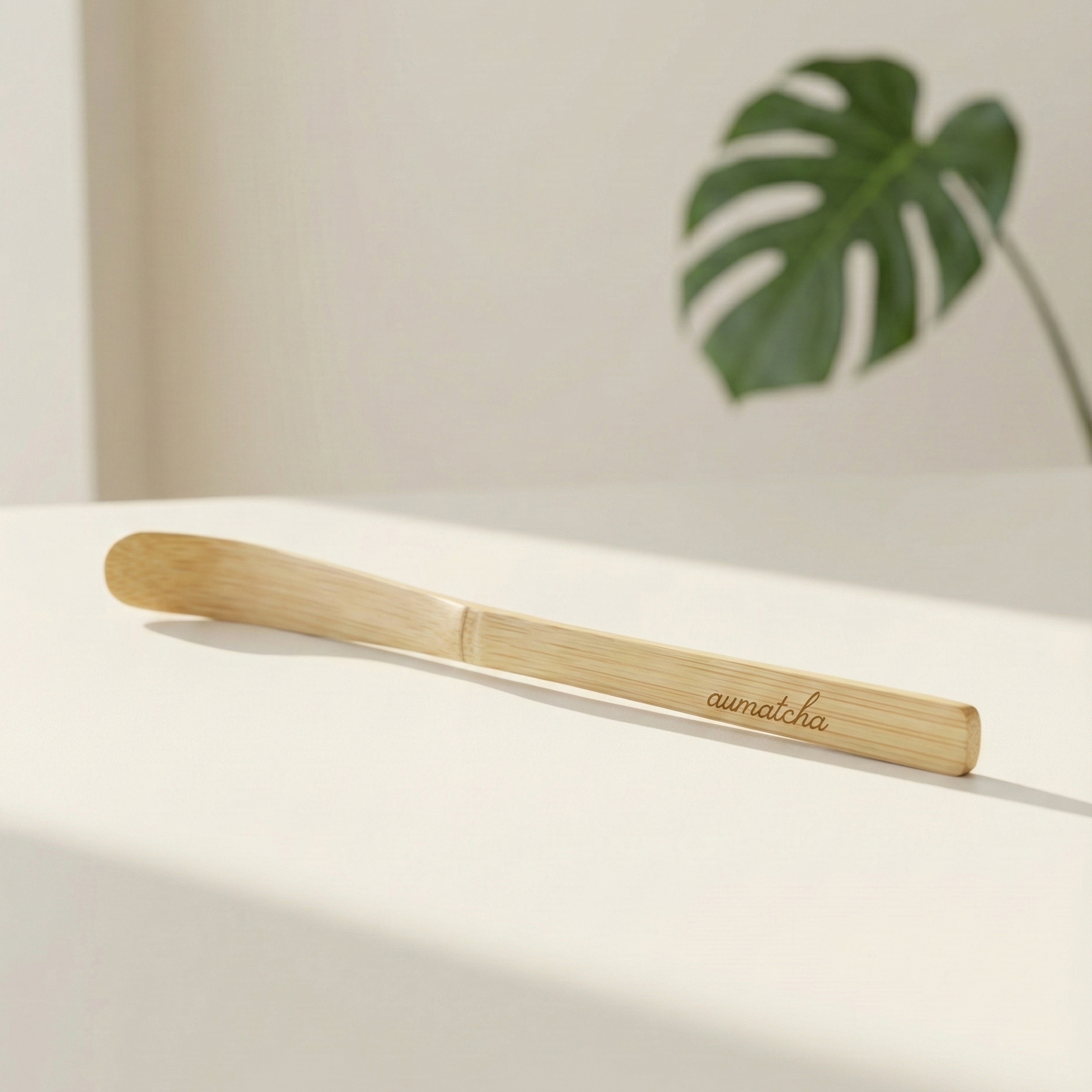 Bamboo tea spoon with 'aumatcha' branding on a light surface with a plant in the background
