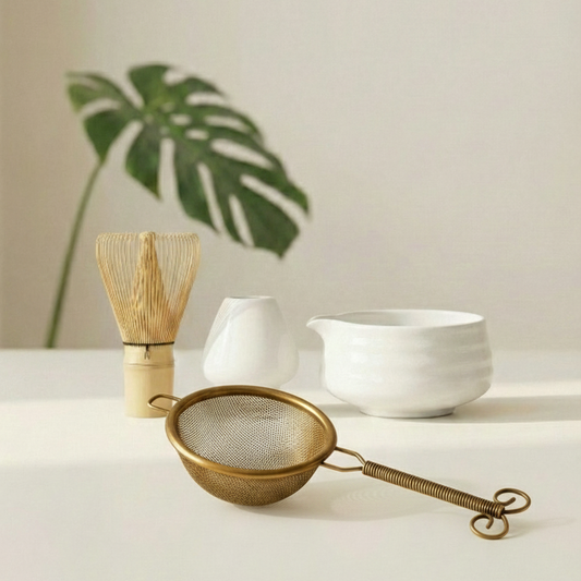 Matcha tea-making tools including a wooden whisk, white bowl, and metal sieve on a light surface with a plant in the background.